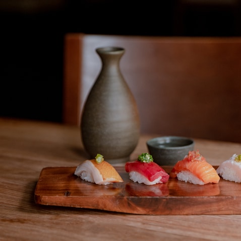 Four different sushi menu items served on a wooden plate next to a tokkuri and ochoko.